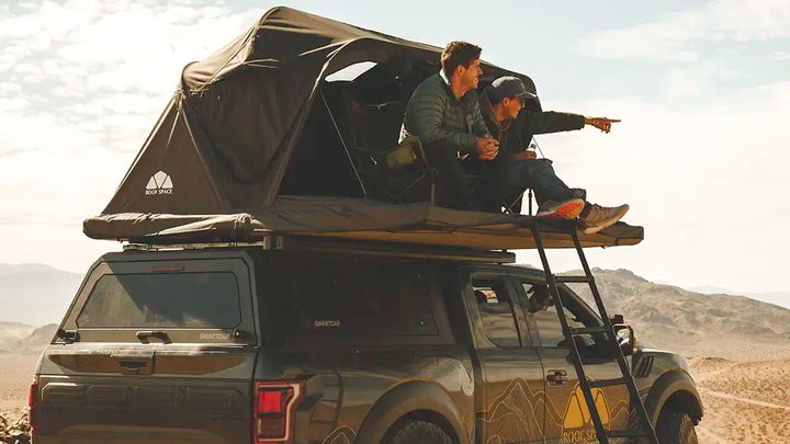 Two people sitting on a Roof Space 4 rooftop tent of a vehicle in a desert landscape